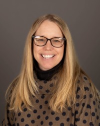 2026-04-10-Kristen-Kostlielney-01-200x250 Headshot of Kristen Kostlienley wearing a polka dot shirt and glasses against a grey background.
