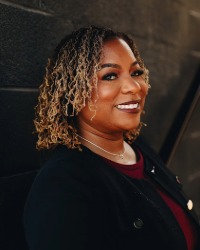 2026-04-10-Tanisha-Bolden-01-200x250 Headshot of Tanisha Bolden wearing a black cardigan against a dark grey brick wall.
