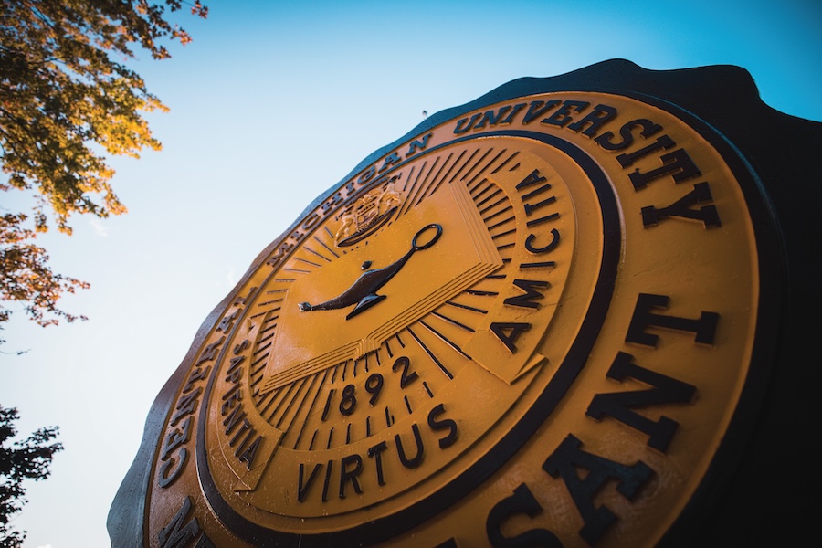 A close up of the CMU Seal, maroon and gold against a blue sky and green trees.