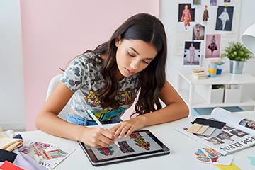 a woman sitting at a table and using a digital tablet to draw fashion images