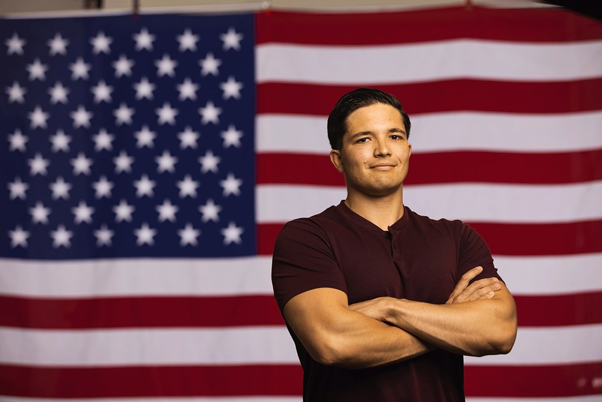 CMU military veteran alumni wearing a short sleeve polo shirt stands in front of an American flag with his arms folded.