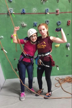 Chloe Straub and Sarah Spearing in Adventure Seminars with rock climbing gear on and the rock climbing wall behind them.