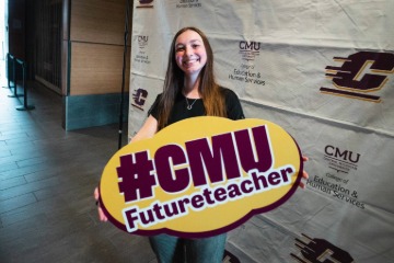 A student holding a sign that says hashtag CMUFutureTeacher with a white CMU banner behind them.