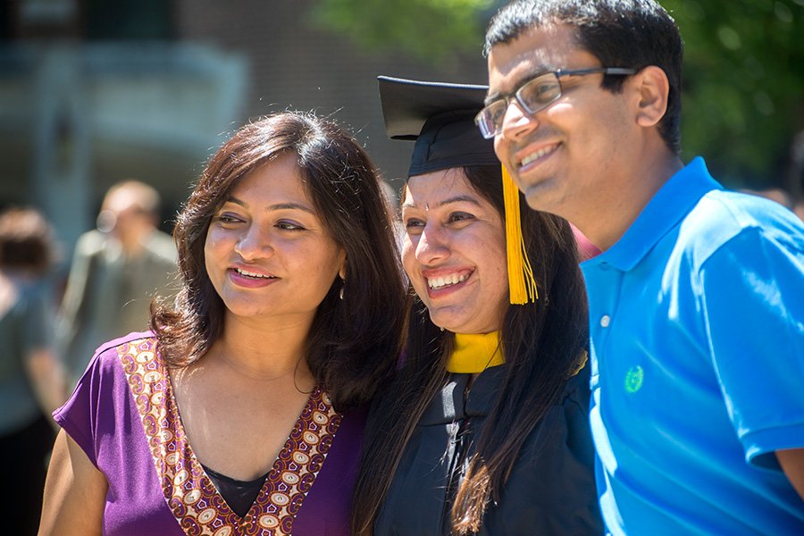 Two women with dark hair, one wearing a graduation cap and gown, stand with a man wearing glasses and a blue polo shirt.