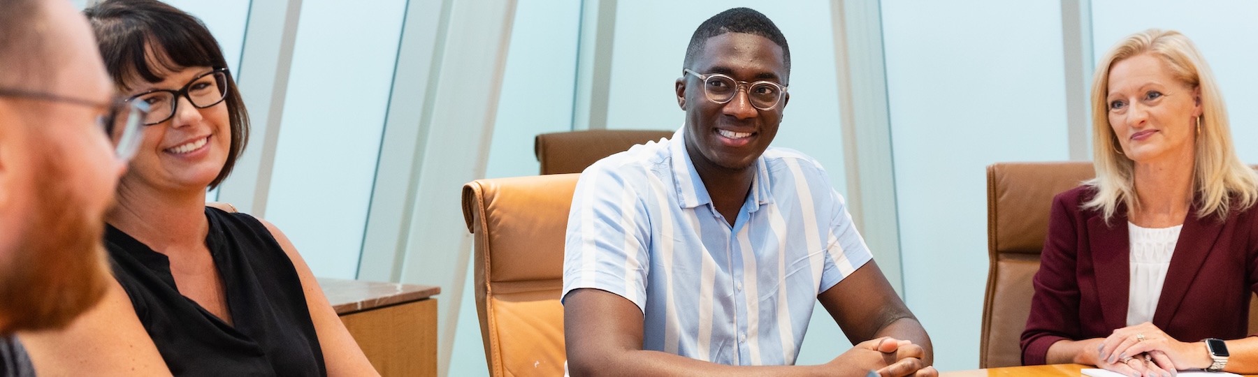 Four individuals are sitting around a table and sitting in tan chairs while smiling and talking with one another.