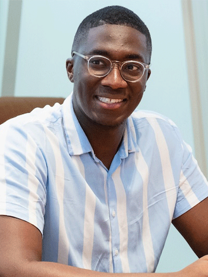 A close up of a student whom is wearing a blue tshirt and glasses, sitting in a tan chair while smiling and talking with others near.