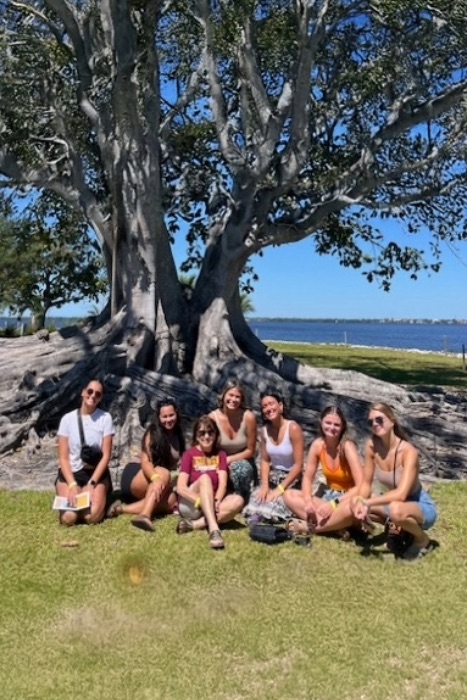 20251119-Immokalee-Study-Abroad-01-467x700 Students sitting in the grass in front of a very large tree in Florida.