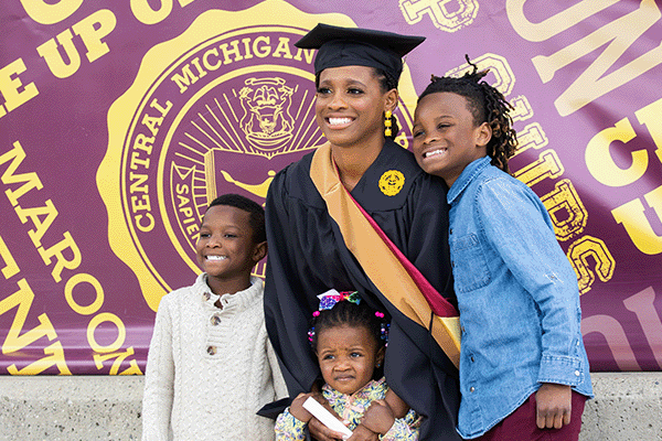 2022 Graduate Commencement Spring On graduate commencement day an adult female wearing a cap and gown poses with her three proud children.