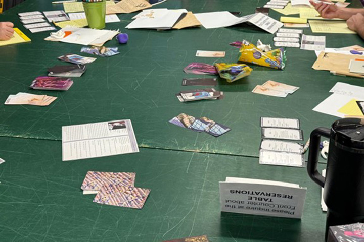Image of board game cards and papers laid out on a green table while two individuals write on notepads.
