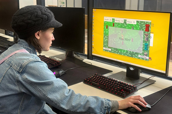 A student sits at a table with a computer monitor to play a video game.
