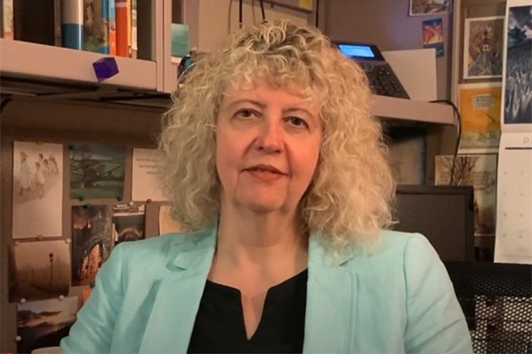 Anne Alton wearing a green suit coat and black shirt is seated in her office with books and images behind her