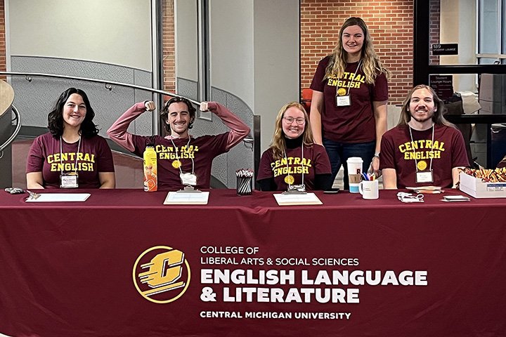 Five CMU students wearing Central English t-shirts sit and stand at a registration table with an English Department table cloth.
