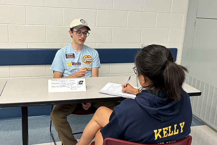 An individual wearing a blue t-shirt and hat answers a question while seated across the table from an individual wearing a dark blue sweatshirt who is writing in a notebook.