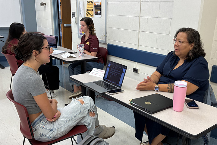 A student wearing a grey t-shirt and jeans seated at a table with her computer in front of her interviews a faculty member wearing a dark blue shirt seated across the table from her.
