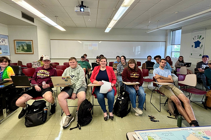 Melissa Stanley (center) wearing a red suit jacket sits at a desk in her classroom with students seated at desks next to her and behind her.