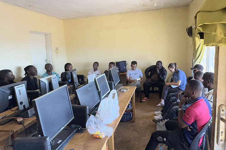 Prakash Adhikari and Molly Dunn hold a discussion with Global Classroom students in Malawi while seated in chairs around the edge of a classroom with rows of tables and computers between them.