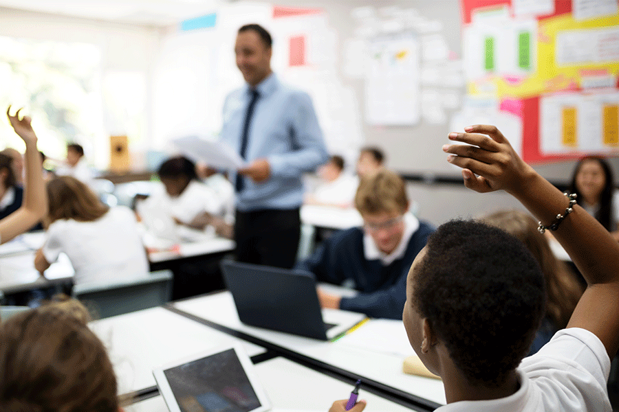 Teacher standing in the middle of a classroom with students sitting at desks with their hands raised to answer a question.