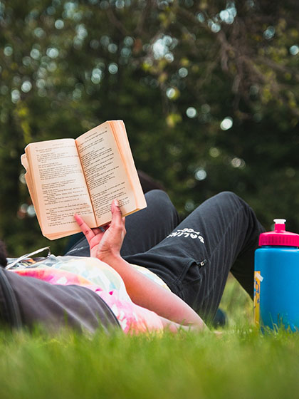 English literature student wearing black sweatpants and a tie-dye shirt lays back on the grass while holding a book up and reading during an outdoor class.