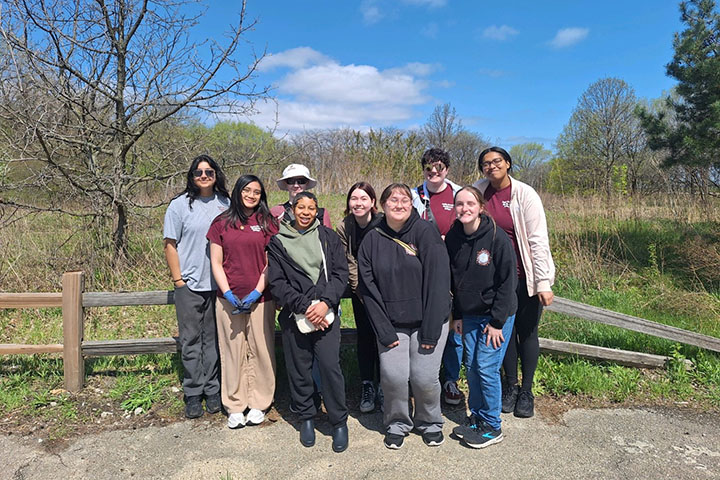 Individuals stand together in front of a wooden fence with grass and brush behind them.
