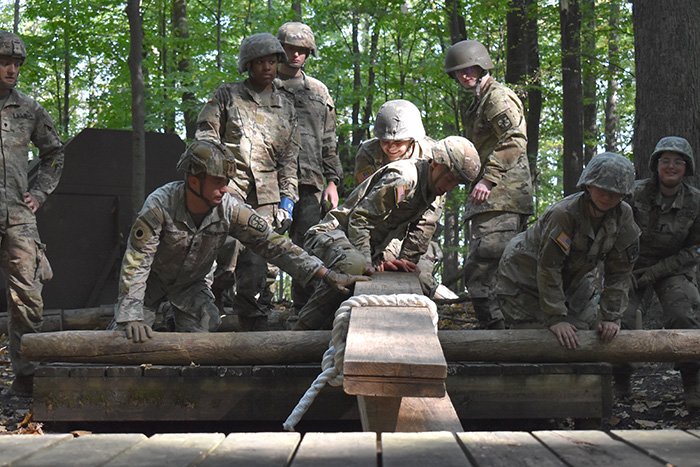 Military Science cadets wearing military uniforms balance wooden beams in the wooded Field Leaders Reaction Course