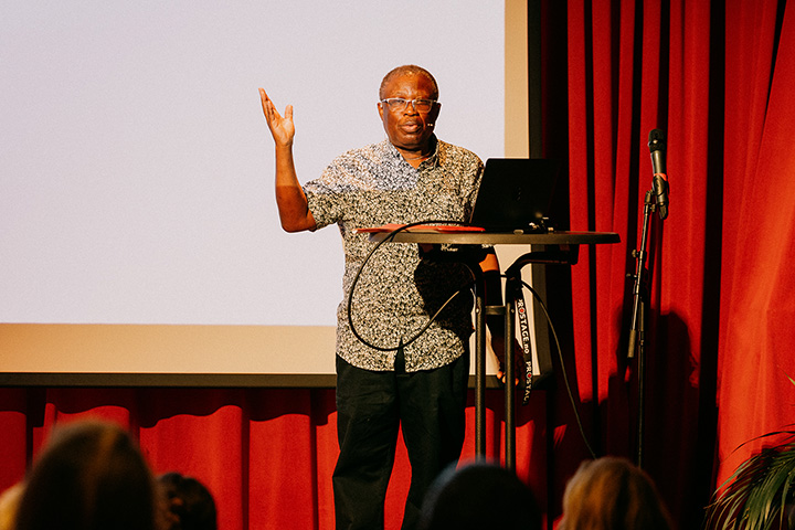 Mensah Adinkrah stands on stage next to a podium and in front of a large screen and red curtain during Heksefest in Norway
