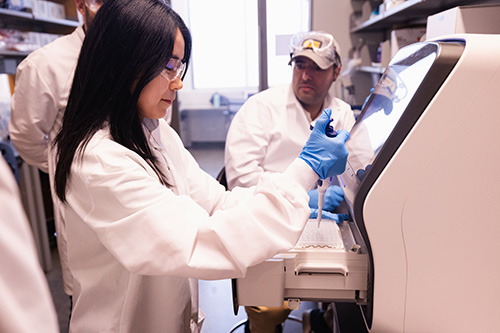 A student in a white lab coat and blue gloves uses a pipette while working with a lab machine, as other researchers observe in a laboratory setting.