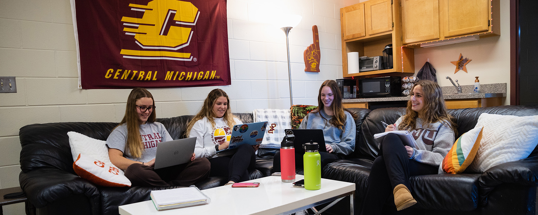 A group of students in a cozy residence hall room sitting on couches with laptops having a discussion.