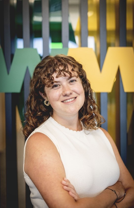 Ava Brewer in a Maroon Cap and Gown standing underneath a brick archway that says Central Michigan University.