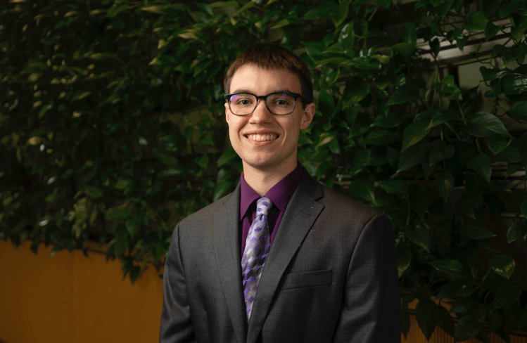 Eric Urbaniak in a maroon cap and gown standing in front of the CMU seal in front of Warriner Hall.