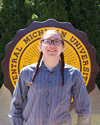20240516_CS-MasonSquillets-Peterson 200x250 Mason Squillets'Peterson in a striped long sleeve shirt and glasses, standing in front of the CMU seal, smiling at the camera.
