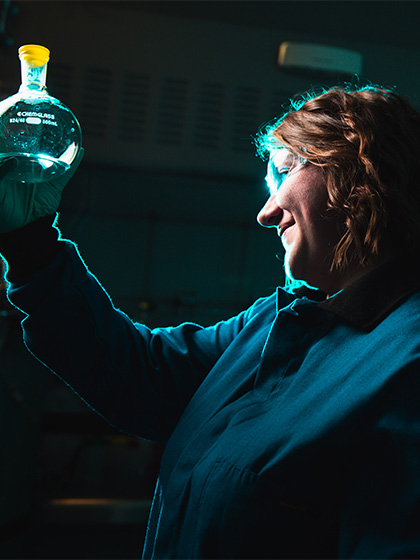 A student in safety glasses and a blue lab coat holding up a container with a liquid in it.