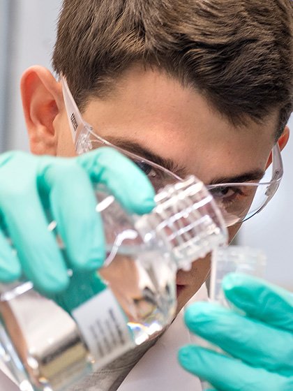 A student wearing goggles and gloves pouring liquid from 1 container into another.