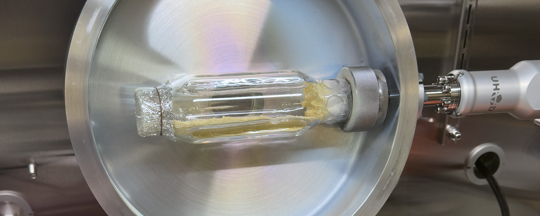 A close-up view of a sealed glass vial containing yellow powder, secured inside a high-vacuum chamber.