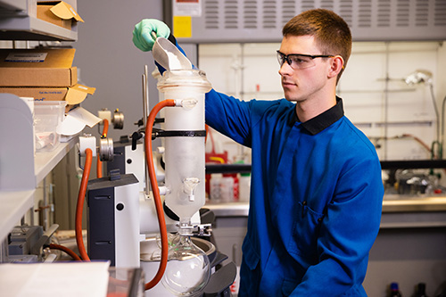 A student wearing safety goggles and a blue lab coat carefully pouring a substance into the glass apparatus.