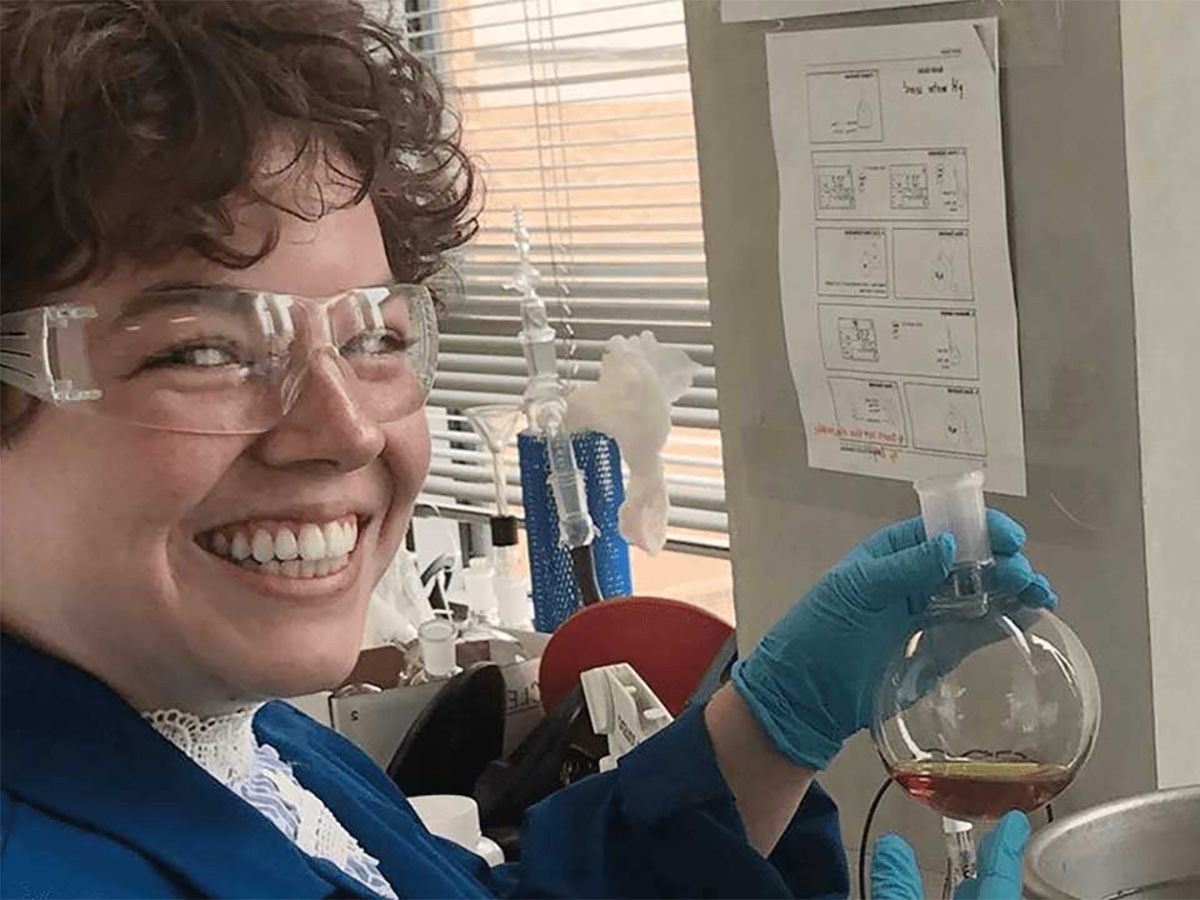 A woman in the blue lab coat with safety goggles on holding a beaker containing a dark liquid.
