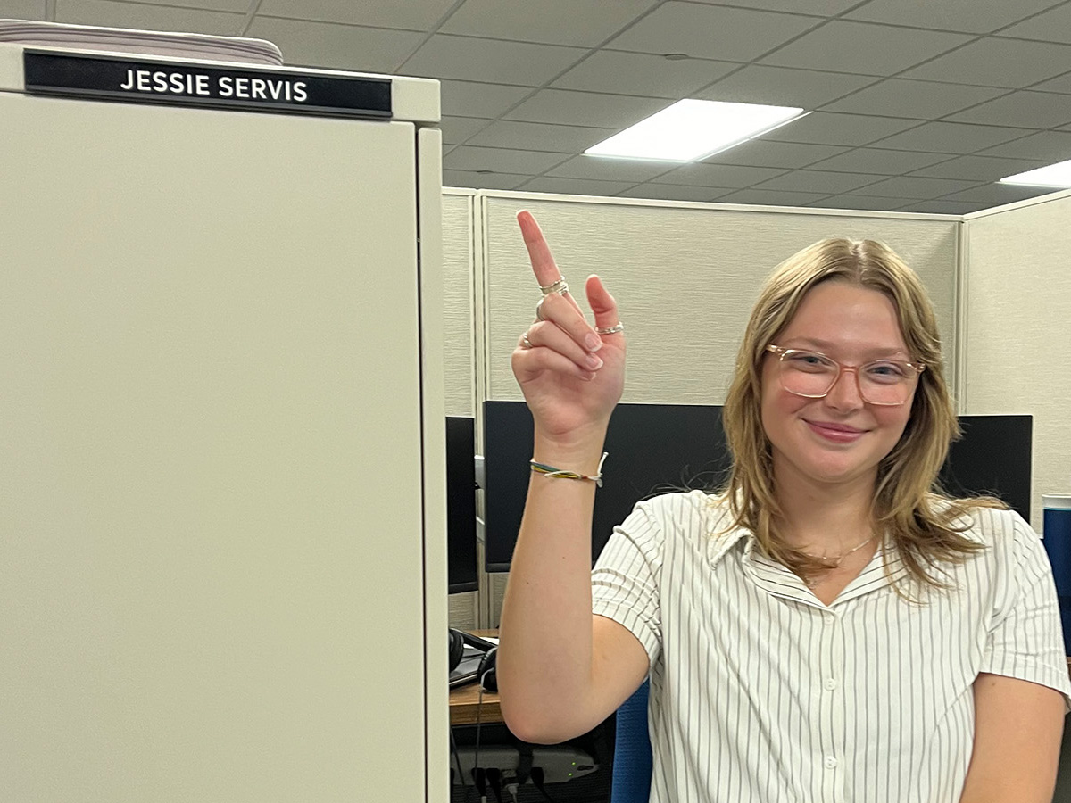 A woman in an office cubicle pointer at a name plate that says Jessie Servis