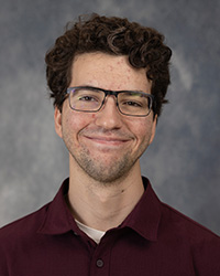 A man with dark hair and a maroon polo shirt wearing glasses and smiling.