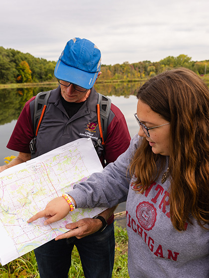 A student and professor stand near a calm lake, studying a topographic map together.