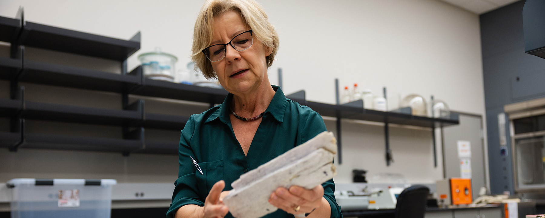A woman in a dark green shirt and glasses examines a rectangular sample in a laboratory setting.