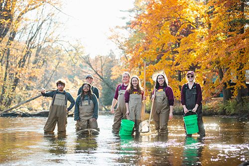 A group of people with waders on standing in a river holding buckets.