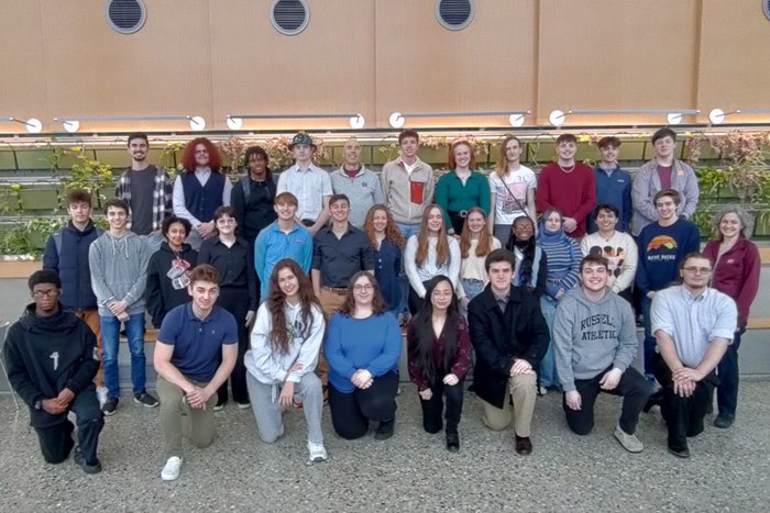 Students and faculty grouped together in front of a green wall in the Biosciences Building at CMU.