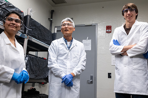 Three individuals in lab coats and safety goggles stand in front of a wall of fish tanks.