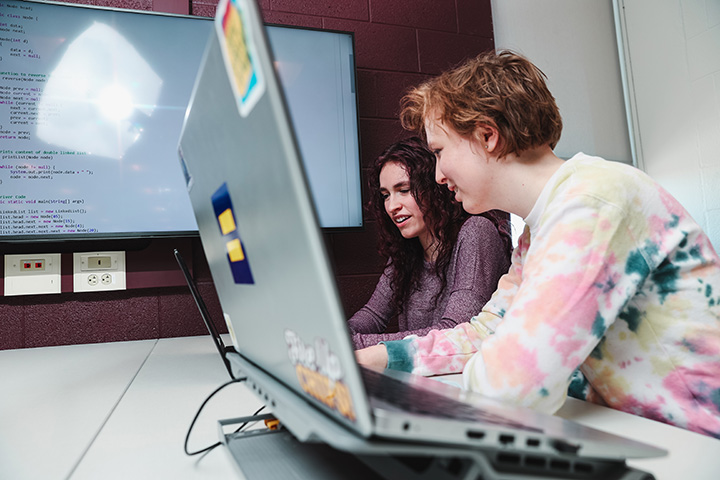 Two computer science students viewing code on their laptops and on a larger screen on the adjacent wall.