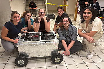 A group of students kneeling around a silver container with wheels and a solar panel on top.