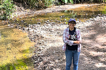 A woman in jeans, checkered shirt and hat standing on a river bed.
