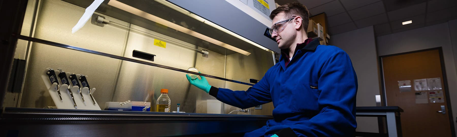 A male student in a blue lab coat and safety goggles holds a petri dish under a hood in a research lab on campus.