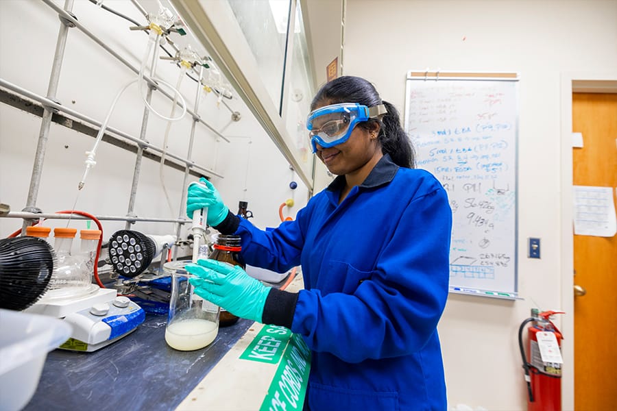 A biochemistry student holds a beaker and pipette while working in a lab on campus.