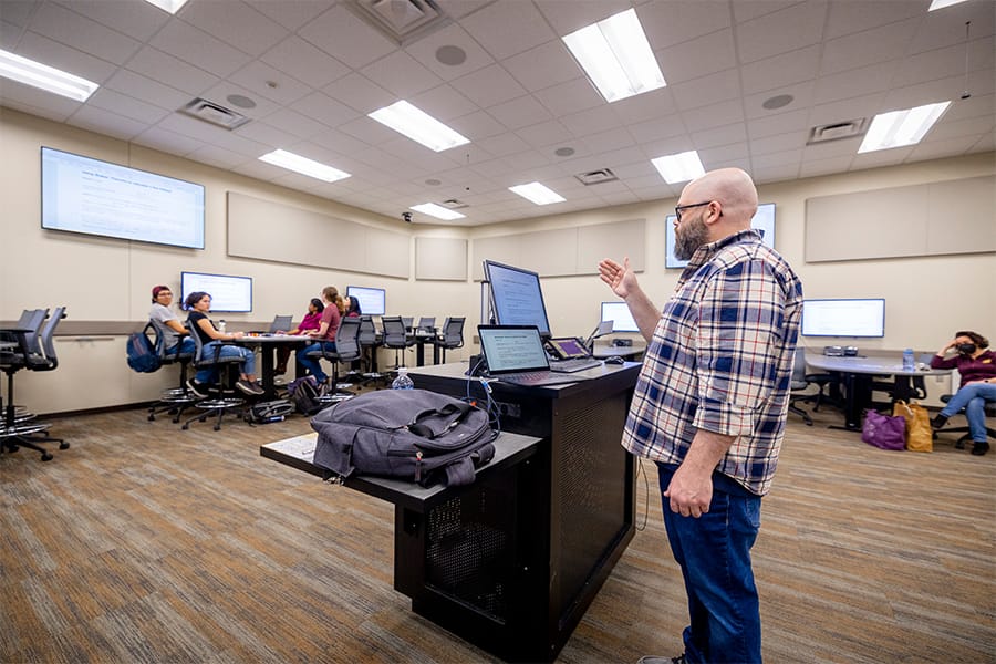 A man in a plaid shirt stands in front of a group of mathematics students sitting at a table together.