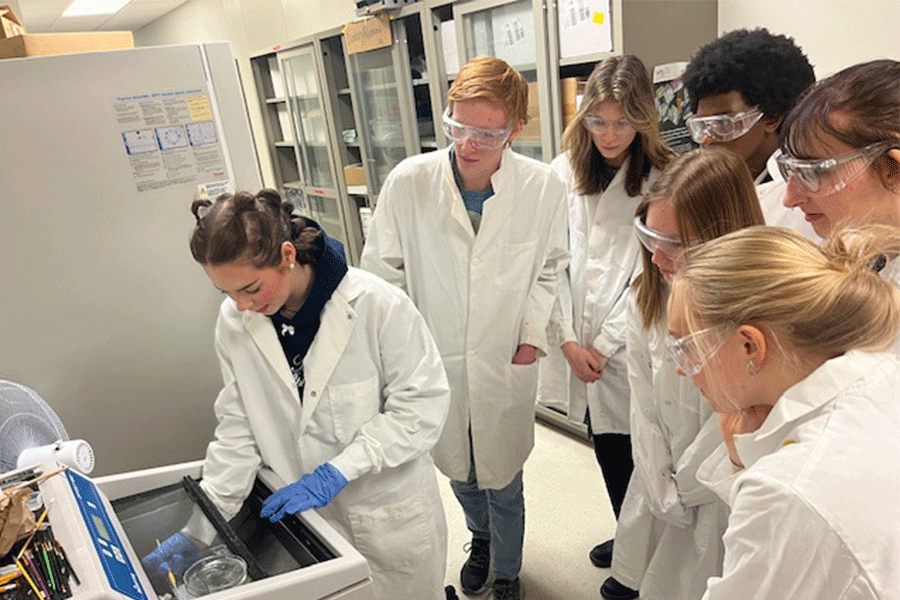 A group of students in white lab coats stand around a glass beaker as one student works in a machine with protective googles on.