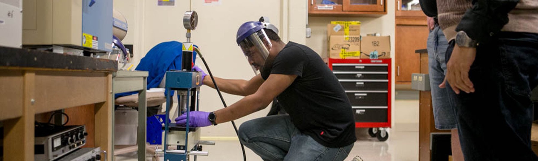 A student wearing a face shield and plastic gloves works in a physics lab on campus.